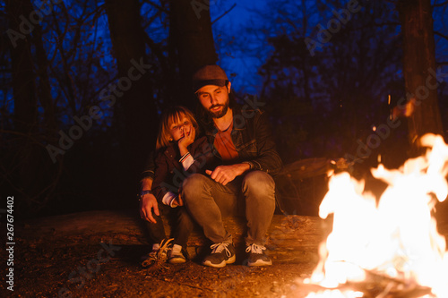 Happy father and his little son sitting together on the logs in front of a fire in a hike in the forest at the night.
