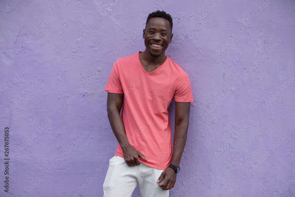 Smiling African American man model posing in empty living coral color t-shirt standing against violet wall background Smiling African American man model posing in empty living coral color t-shirt standing against violet wall background