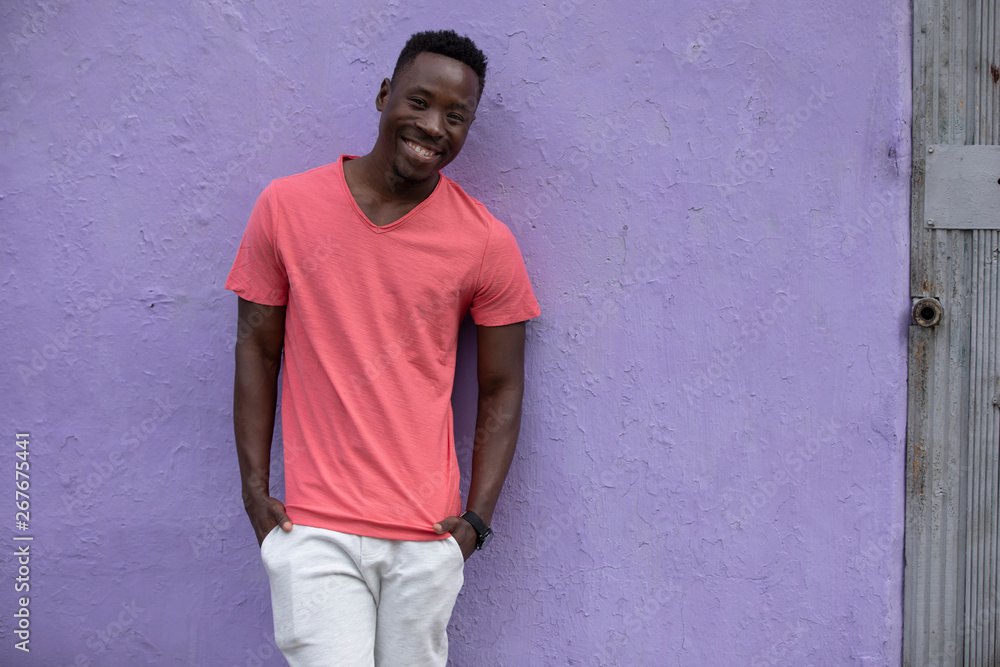 Smiling African American man model posing in empty living coral color t-shirt standing against violet wall background Smiling African American man model posing in empty living coral color t-shirt standing against violet wall background