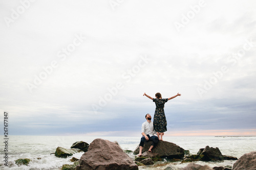 happy couple in love on the beach. man sitting on the rocks and woman standing with open hands