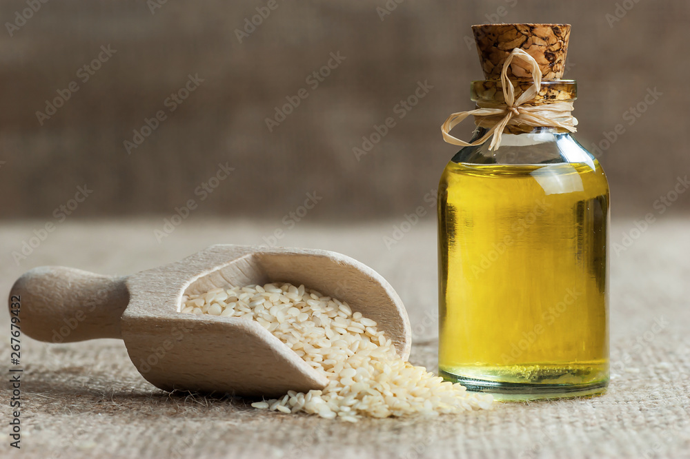 Φωτογραφία Glass bottle of sesame oil and raw sesame seeds in wooden shovel or spoon on burlap sack