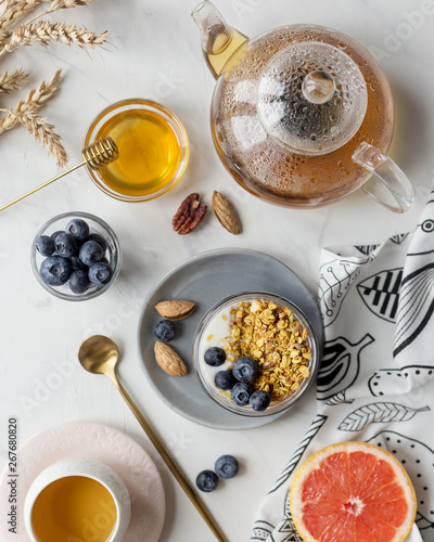 Granola in jar with blueberries with tea, honey and grapefruit on white marble background