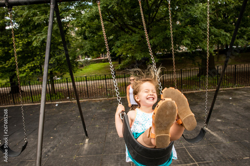 A toddler aged girl swinging and having fun outdoors at a park with her mother.