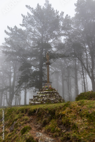 Traditional Galician stone crossroads surrounded by trees and vegetation on a foggy day. Cruceiro.
