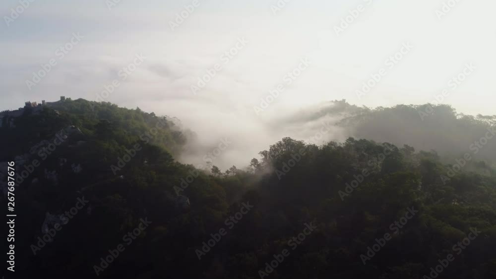 Aerial view Castelo dos Mouros on top of mountain in sunny fog morning. Drone flying above forested hills at the distance walls of medieval fortress on peak. Portugal, Sintra Aerial view Castelo dos Mouros on top of mountain in sunny fog morning. Drone flying above forested hills at the distance walls of medieval fortress on peak. Portugal, Sintra