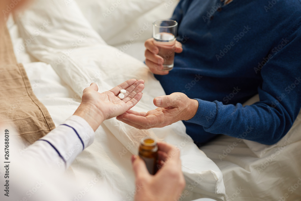 Closeup of unrecognizable senior man lying on bed and taking pills with caring wife or nurse , copy space