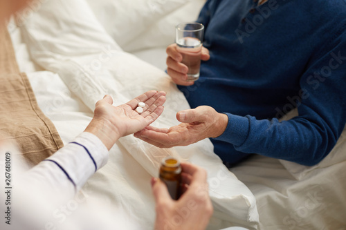 Closeup of unrecognizable senior man lying on bed and taking pills with caring wife or nurse   copy space
