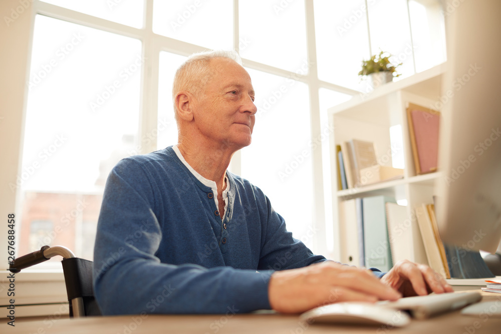 Low angle portrait of smiling senior man sitting in wheelchair using computer at home, copy space