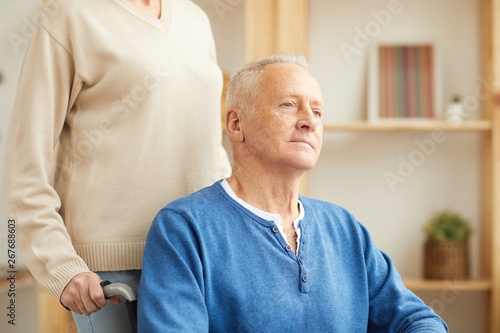 Portrait of pensive senior man in wheelchair looking away led by wife of caregiver, copy space