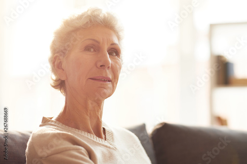 Head and shoulders portrait of mature woman looking at camera while posing at home lit by sunlight, copy space