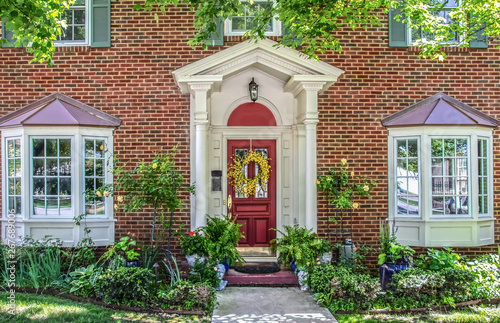 Entrance with columns and bay windows of beautiful two-story brick house with pots of flowers and yellow climbing roses and headplanters and ferns frame by Maple tree limbs