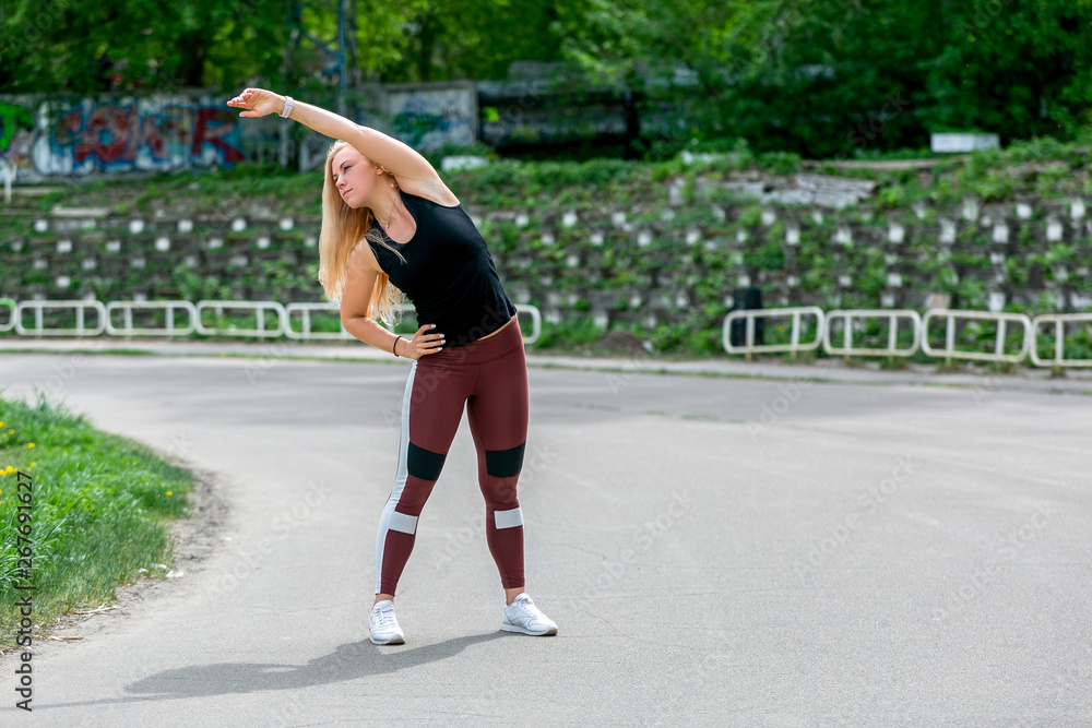 Fitness lifestyle. Young woman warming up before training doing exercises to stretch her muscles and joints. Sporty young blonde girl on a sunny day at the stadium. Healthy life concept