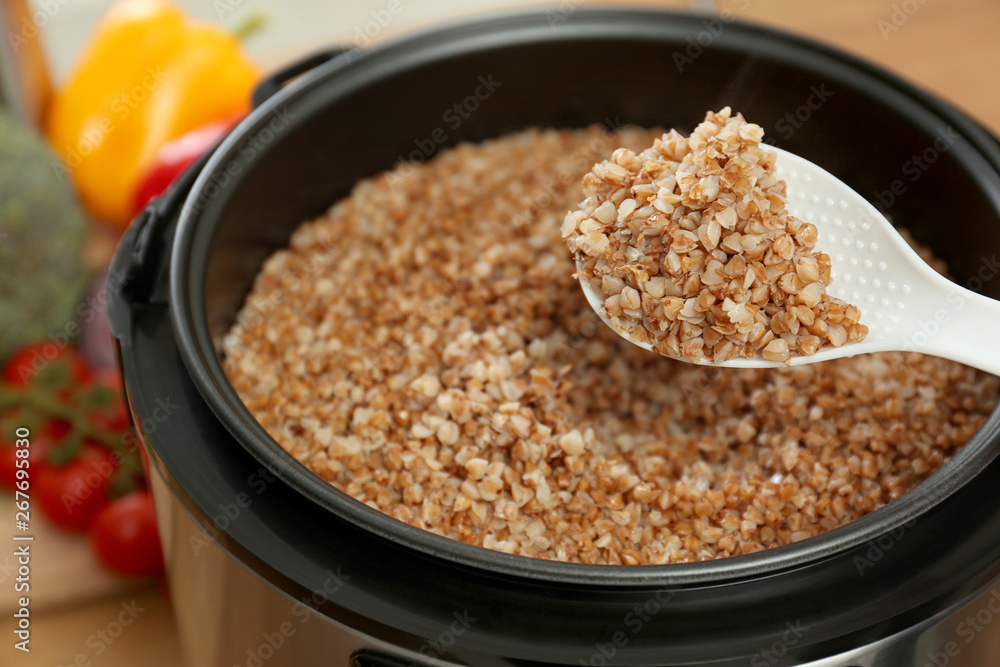 Spoon with delicious buckwheat over multi cooker in kitchen, closeup Spoon with delicious buckwheat over multi cooker in kitchen, closeup