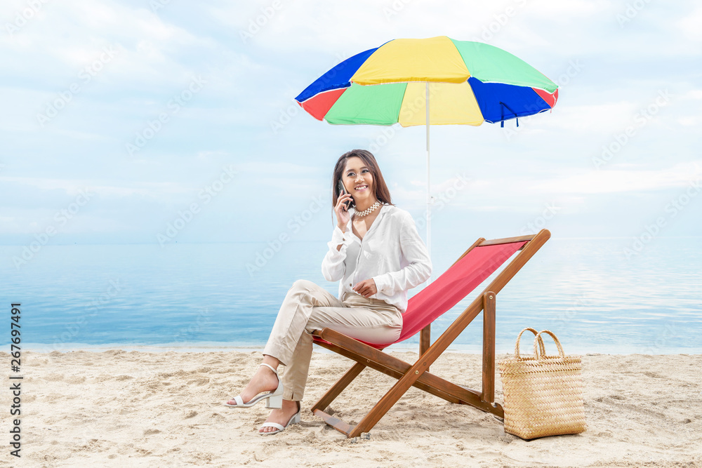 Asian business woman talking on mobile phone while sitting sitting in the beach chair with colorful umbrella on beach