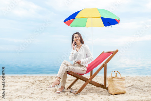 Asian business woman talking on mobile phone while sitting sitting in the beach chair with colorful umbrella on beach © Leo Lintang