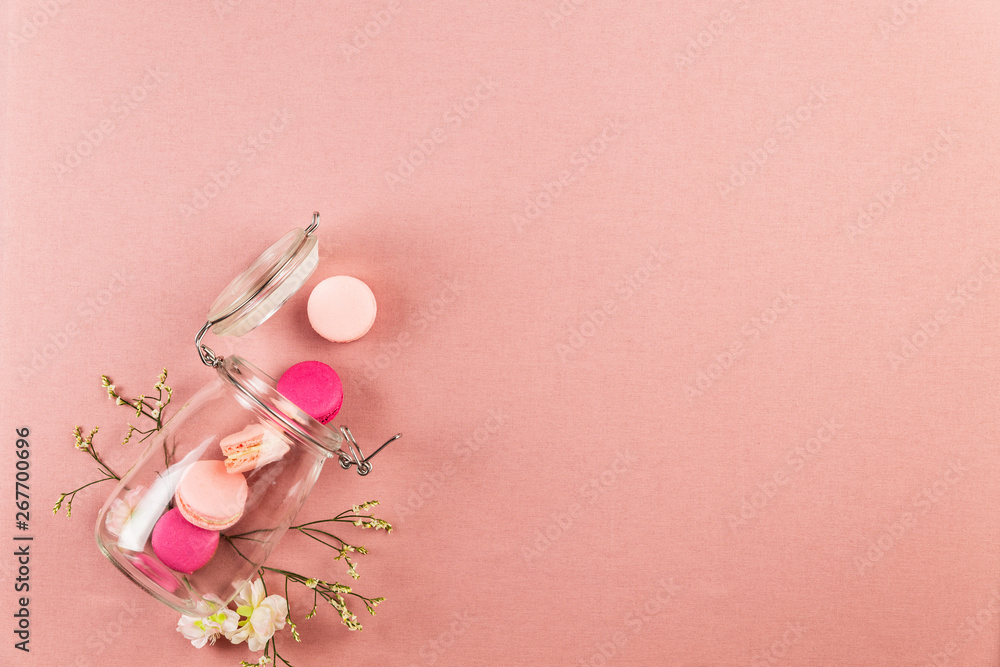 Pink french macarons or macaroons, falling out of a glass jar with white flowers over a pink tablecloth background with copy space.