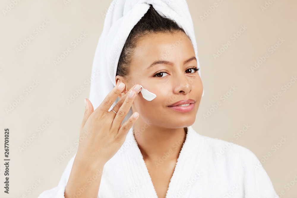 Head and shoulders portrait of smiling Mixed-Race woman applying face cream and looking at camera during morning beauty routine, copy space Head and shoulders portrait of smiling Mixed-Race woman applying face cream and looking at camera during morning beauty routine, copy space