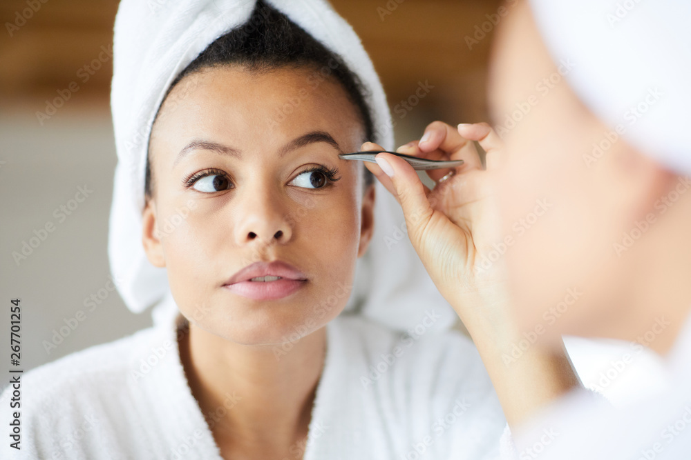 Head and shoulders portrait of  beautiful Mixed-Race woman plucking eyebrows looking in mirror during morning routine, copy space