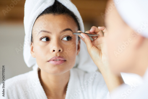 Head and shoulders portrait of  beautiful Mixed-Race woman plucking eyebrows looking in mirror during morning routine, copy space
