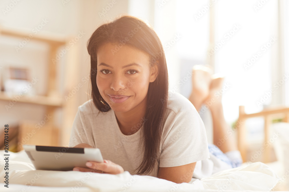 Portrait of beautiful Mixed-Race woman looking at camera while using digital tablet lying on bed lit by sunlight, copy space Portrait of beautiful Mixed-Race woman looking at camera while using digital tablet lying on bed lit by sunlight, copy space