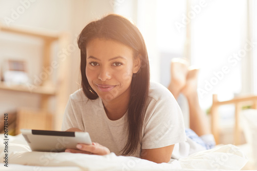 Portrait of beautiful Mixed-Race woman looking at camera while using digital tablet lying on bed lit by sunlight, copy space