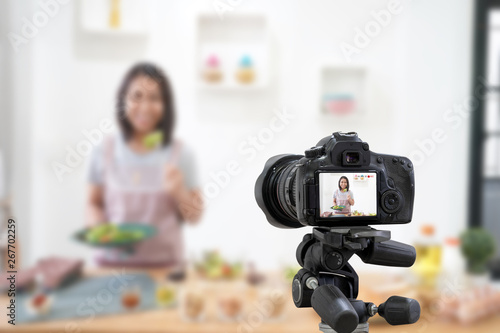 Asian woman making a Vlog video digital camera for her blog cooking in the kitchen room. With holding bowl with green lettuce salad about healthy and nutritious