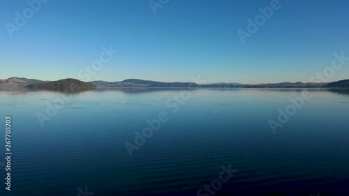 A slowly rising drone shot of Lake Rotorua that pans towards Mokoia Island. Some birds are visible on the lake. photo