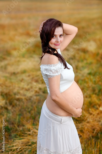 Beautiful pregnant girl in white clothes is standing in a field at sunset and looking into the camera. Close-up. View from above © Маргарита Азимова
