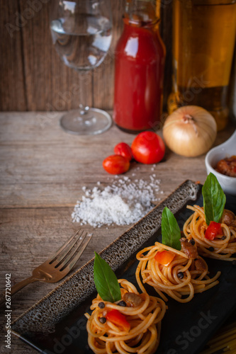 Spaghetti in Tomato sauce fit the pieces on the black plate placed on the wood table there are fork, tomato, sauce, oil, onion, salt and raw spaghetti placed around.