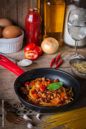 Spaghetti in Tomato sauce on the black pan placed on the wood table there are egg, black pepper, fork, tomato, sauce, oil, onion, salt, chilli and raw spaghetti placed around.