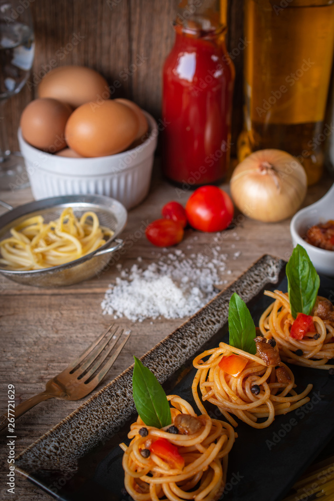 Spaghetti in Tomato sauce fit the pieces on the black plate placed on the wood table there are egg, black pepper, fork, tomato, oil, onion, salt and raw spaghetti placed around. Spaghetti in Tomato sauce fit the pieces on the black plate placed on the wood table there are egg, black pepper, fork, tomato, oil, onion, salt and raw spaghetti placed around.