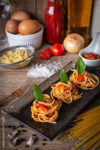 Spaghetti in Tomato sauce fit the pieces on the black plate placed on the wood table there are egg, black pepper, fork, tomato, sauce, oil, onion, salt and raw spaghetti placed around.
