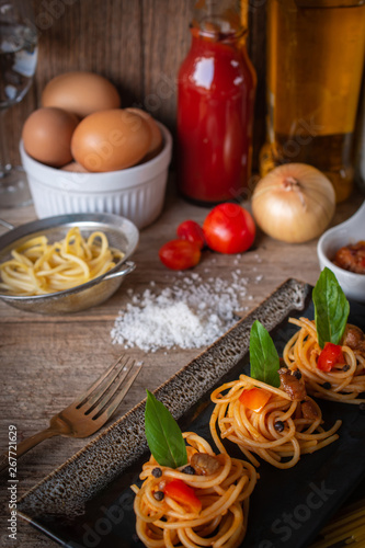 Spaghetti in Tomato sauce fit the pieces on the black plate placed on the wood table there are egg, black pepper, fork, tomato, oil, onion, salt and raw spaghetti placed around.