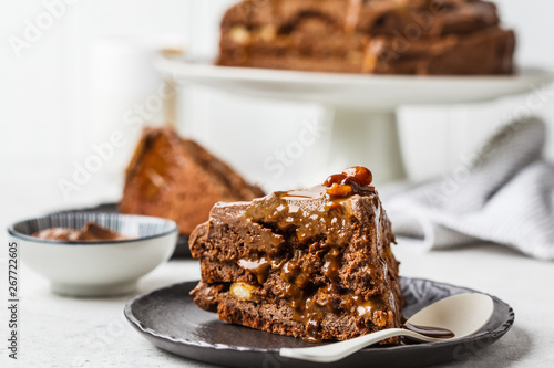 Piece of homemade snickers cake with chocolate cream and caramel on a black saucer, white background.