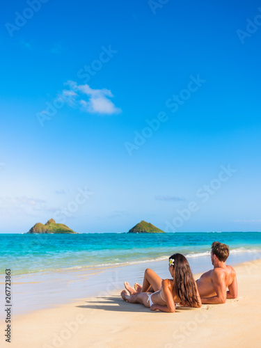 Beach vacation couple relaxing sunbathing on hawaiian tropical beach in Lanikai, Oahu, Hawaii, USA. American people on summer holidays sun tanning lying down on sand.