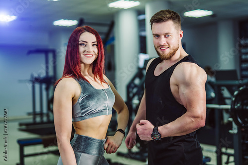 Smiling male and female bodybuilders in sport clothes standing in gym and looking at camera. Young couple with muscular bodies posing together in the fitness center.