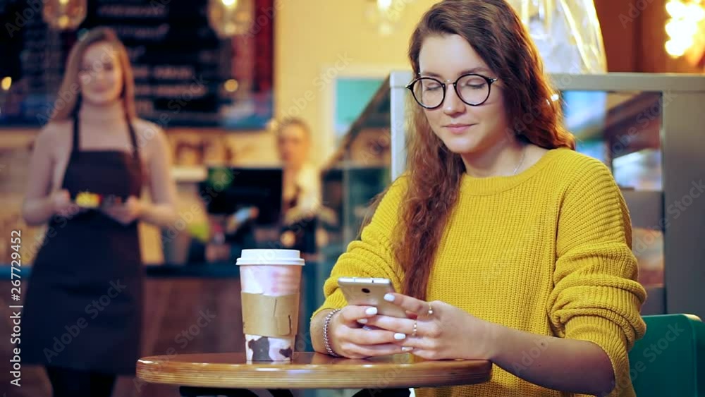 Beautiful girl in glasses sits at a table, she smiles and reads a smartphone message. The waiter comes from a defocused background with dessert Beautiful girl in glasses sits at a table, she smiles and reads a smartphone message. The waiter comes from a defocused background with dessert