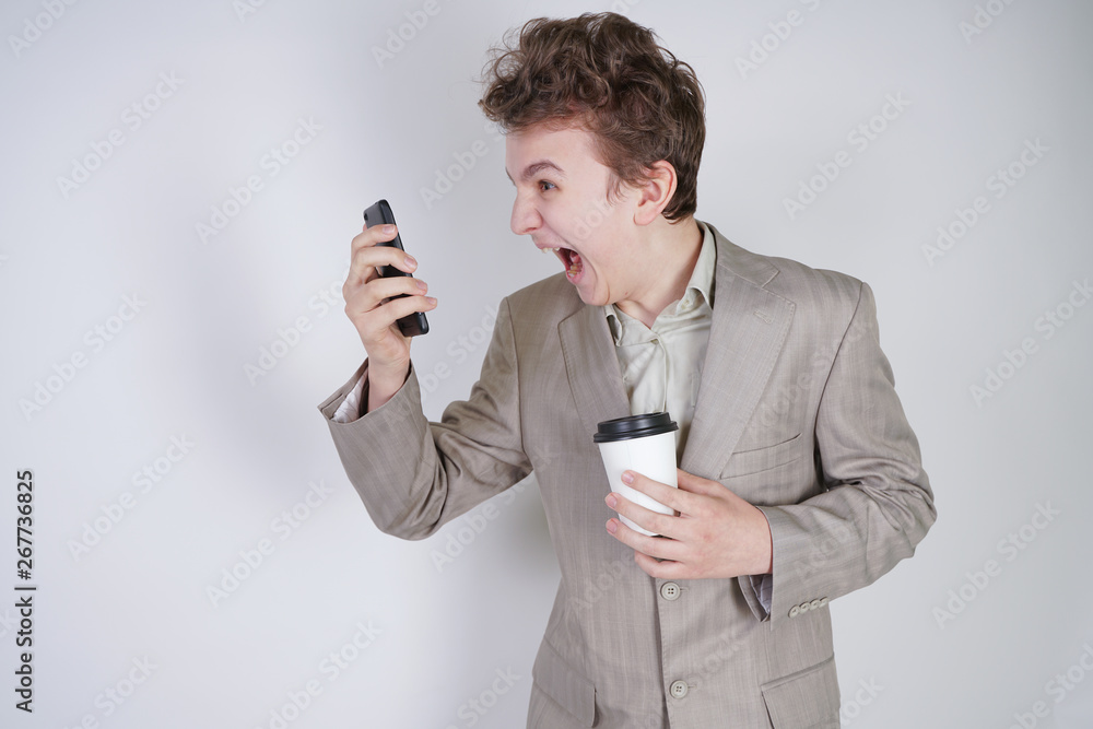 aggressive caucasian teenager screams into the smart phone and holds a paper cup of coffee in his hand. student on edge on a white background in Studio aggressive caucasian teenager screams into the smart phone and holds a paper cup of coffee in his hand. student on edge on a white background in Studio