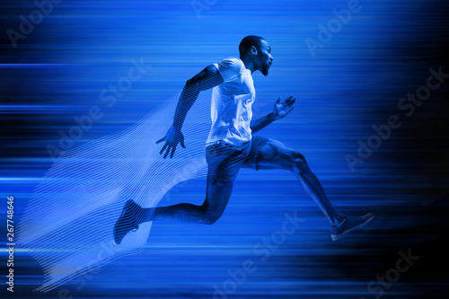 Young african-american man running isolated on blue studio background. One male runner or jogger. Silhouette of jogging athlete with shadows in neon light. Movement or motion. Creative collage. © master1305