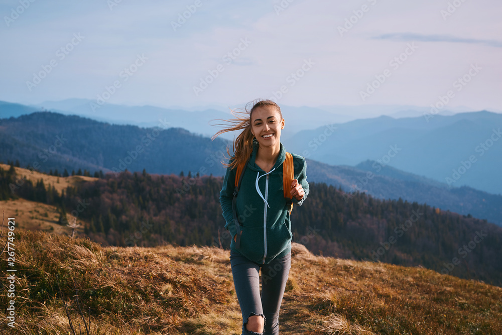 Portrait of a happy woman hiker standing on the slope of mountain ridge against mountains on background. The woman is happy and looking at camera. Travel and active lifestyle concept.