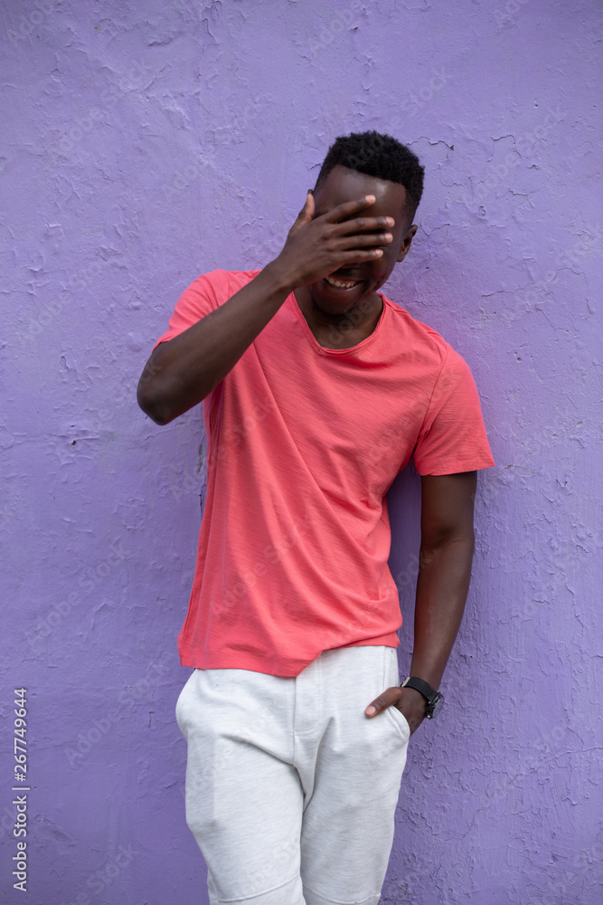 Smiling laughing African American man model posing in empty living coral color t-shirt standing against violet wall background Smiling laughing African American man model posing in empty living coral color t-shirt standing against violet wall background