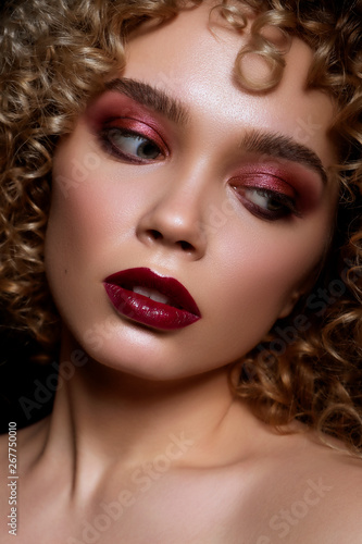 Cute caucasian woman with afro (curls) hairstyle on a dark background. She wears dark evening (or podium) make up with red mascara on her eyes ans lips