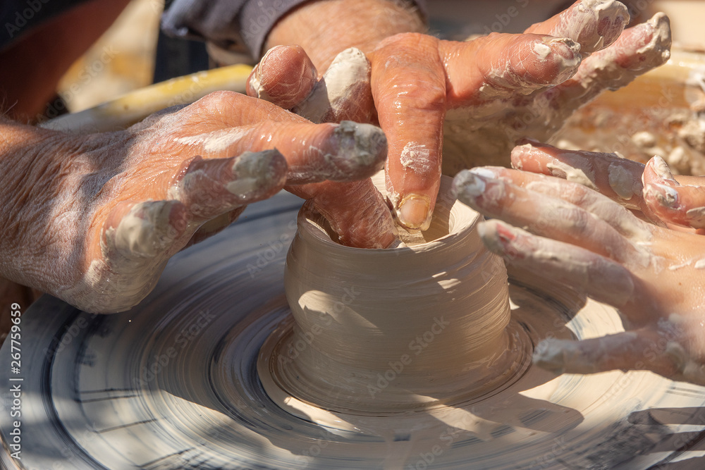 An adult potter helps a young student make a clay pot. Outdoor crafting on potter's wheel. Learning craft An adult potter helps a young student make a clay pot. Outdoor crafting on potter's wheel. Learning craft