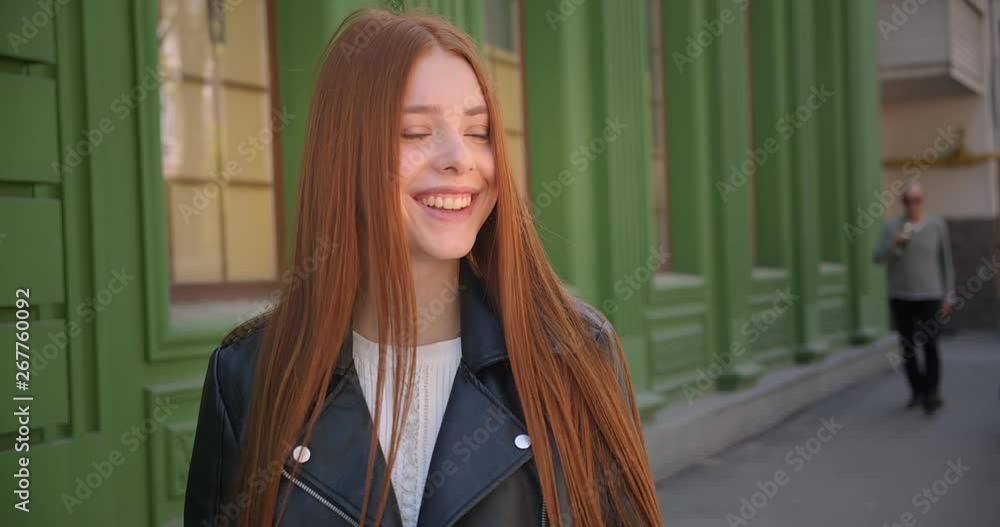 Walking beautiful ginger girl in leather jacket stops near the camera and smiles into it on green house background. Walking beautiful ginger girl in leather jacket stops near the camera and smiles into it on green house background.