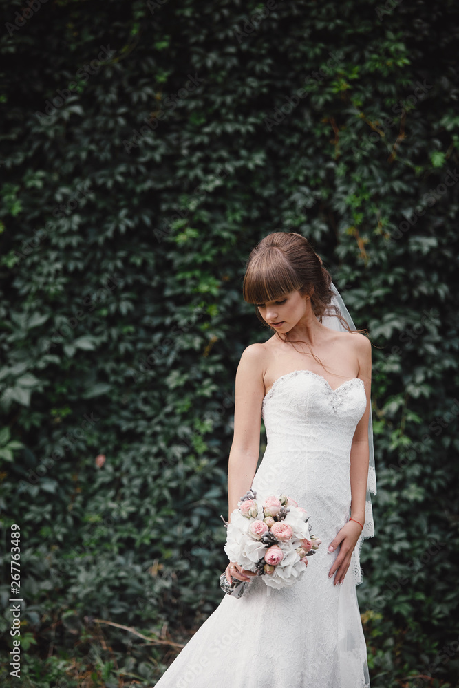 Young beautiful bride in an elegant dress is standing on the field near the forest and holding bouquet of pink flowers and greens with ribbon at nature. Outdoors. After the wedding ceremony. Young beautiful bride in an elegant dress is standing on the field near the forest and holding bouquet of pink flowers and greens with ribbon at nature. Outdoors. After the wedding ceremony.