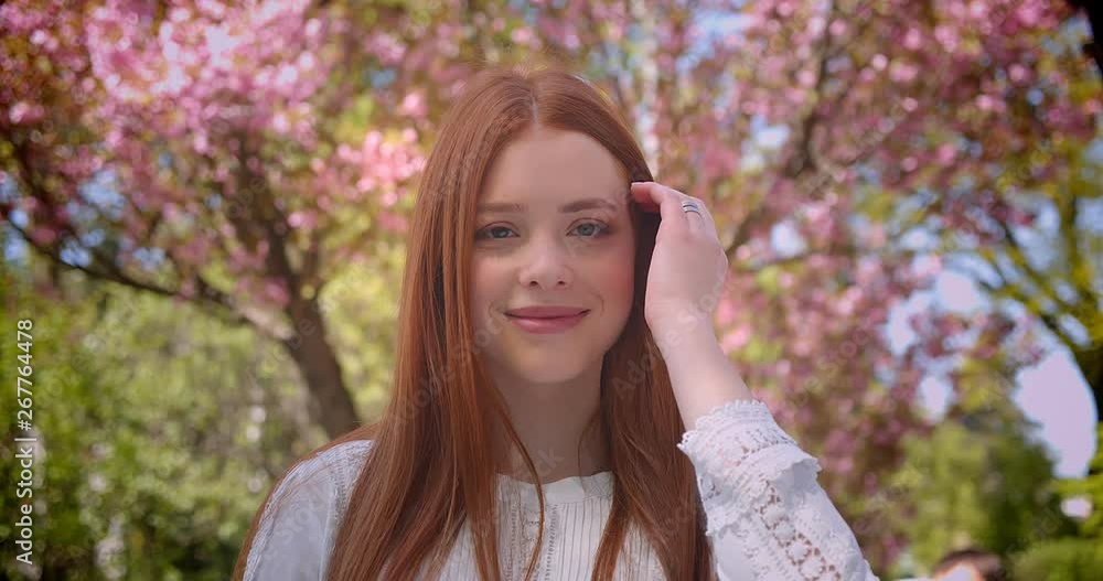 Charming ginger female student in white blouse watching dreamily leftwards and into camera in pink floral garden. Charming ginger female student in white blouse watching dreamily leftwards and into camera in pink floral garden.