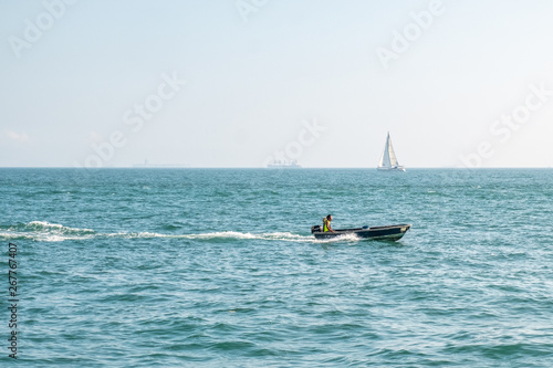 Motor boat moving fast in open sea with foam trace behind. Unrecognizable person in the boat. Sailboat on horizon line. Sunny day. Tropical vacation. Weekend getaway from city. Sport and recreational.