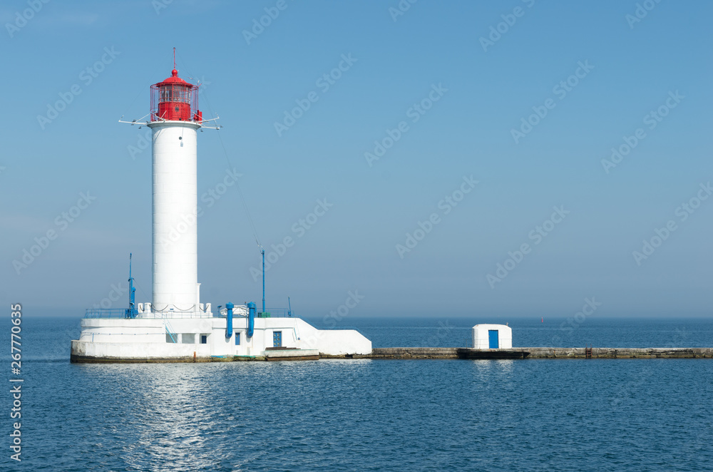 Beautiful big white red lighthouse against the blue calm sea, cloudless sky and clear horizon - a symbol of the safe path for ships. Pharos on the edge of the coast. Seascape of Black sea.