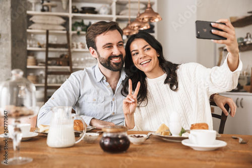 Portrait of lovely european couple taking selfie photo while having breakfast in kitchen at home