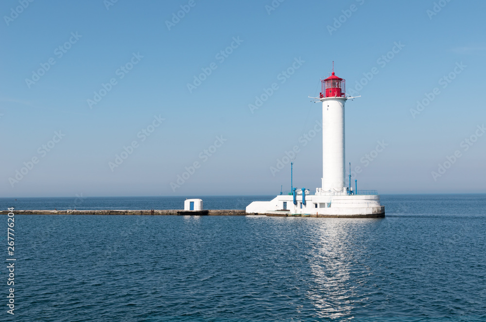 Big white red lighthouse against the blue calm sea, cloudless sky and clear horizon - a symbol of the safe path for ships. Beacon on the edge of the pier. Seascape of Black sea.